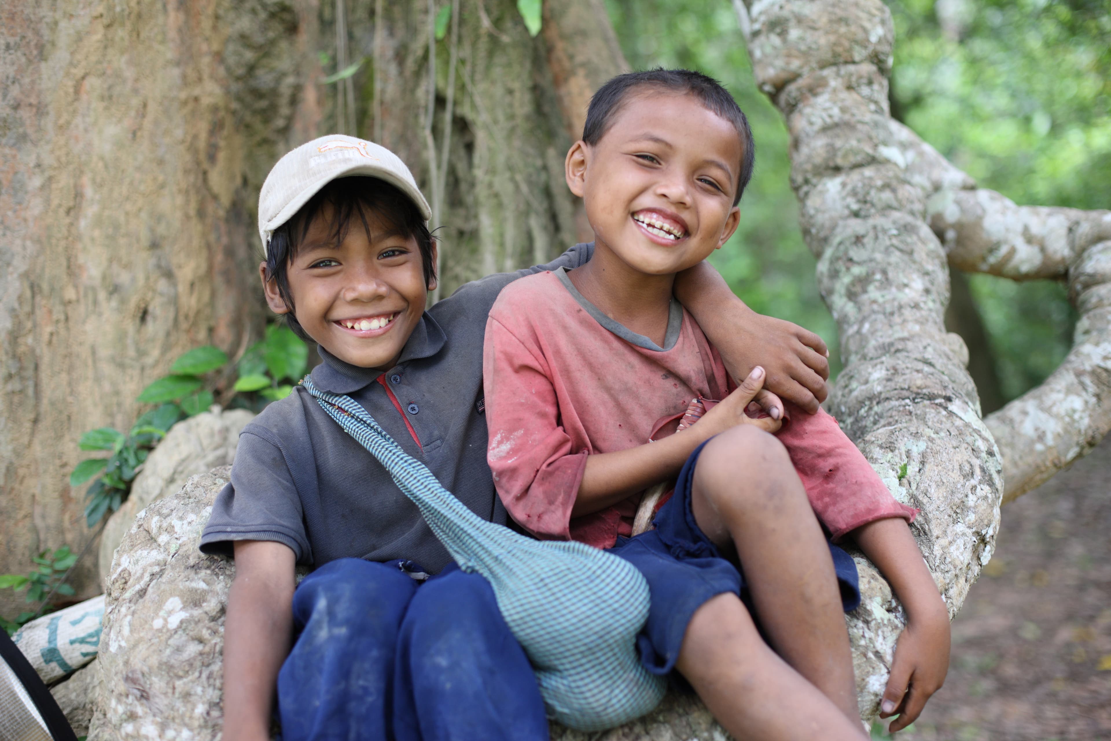 Two boys smiling, sitting together against a tree