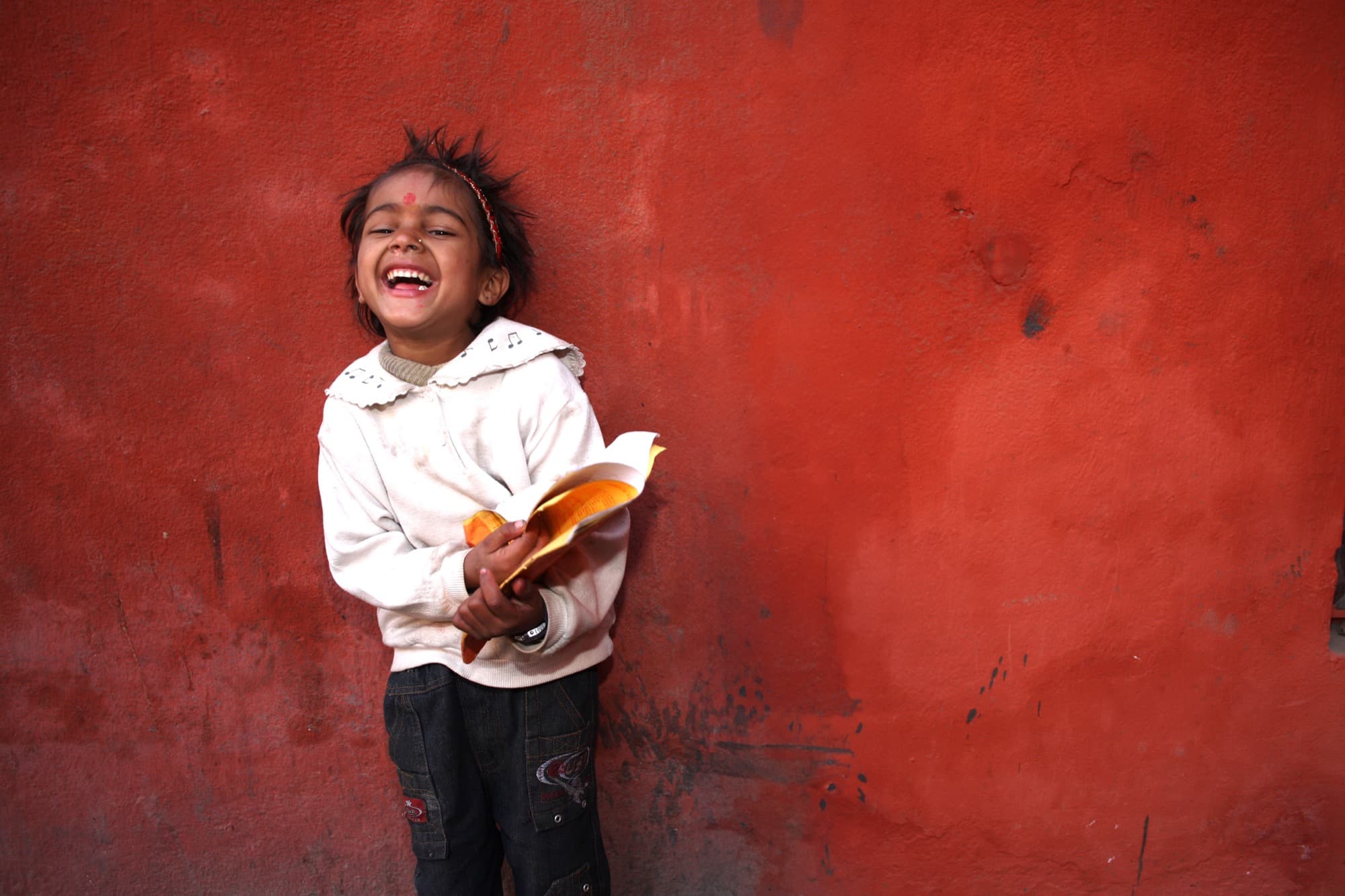 A young girl with a red bindi laughing as she holds a book against a red wall