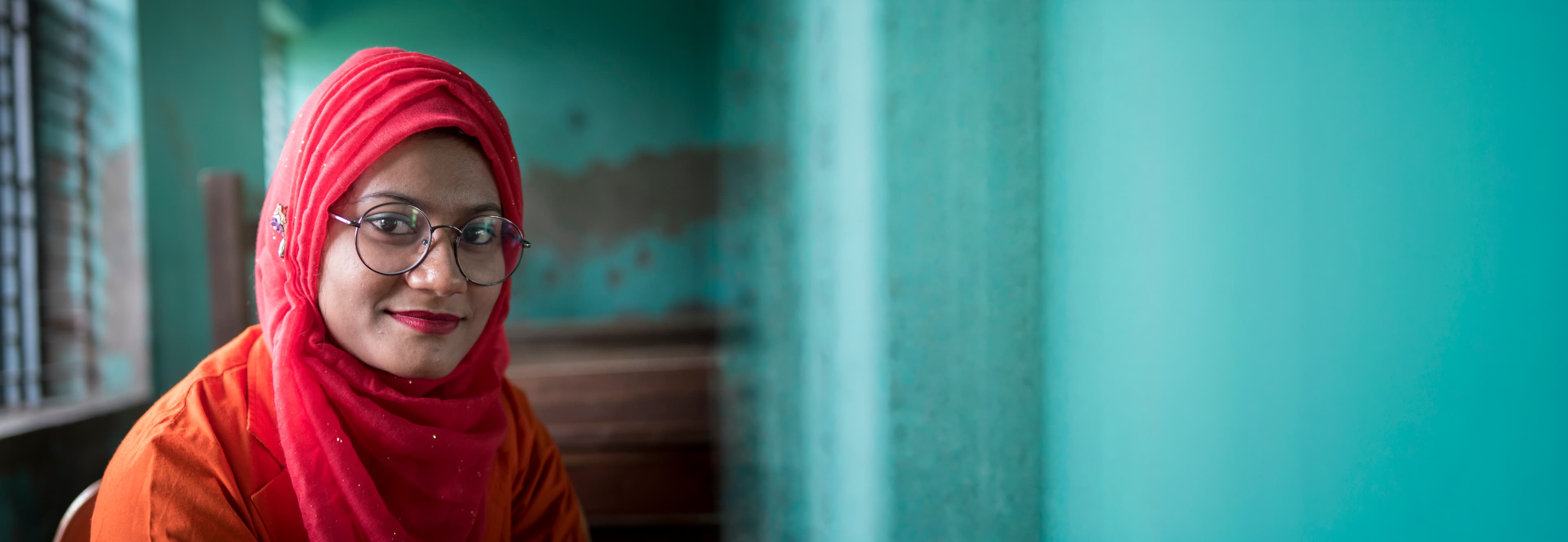 A woman in a red hijab and round glasses, indoor portrait against a teal wall